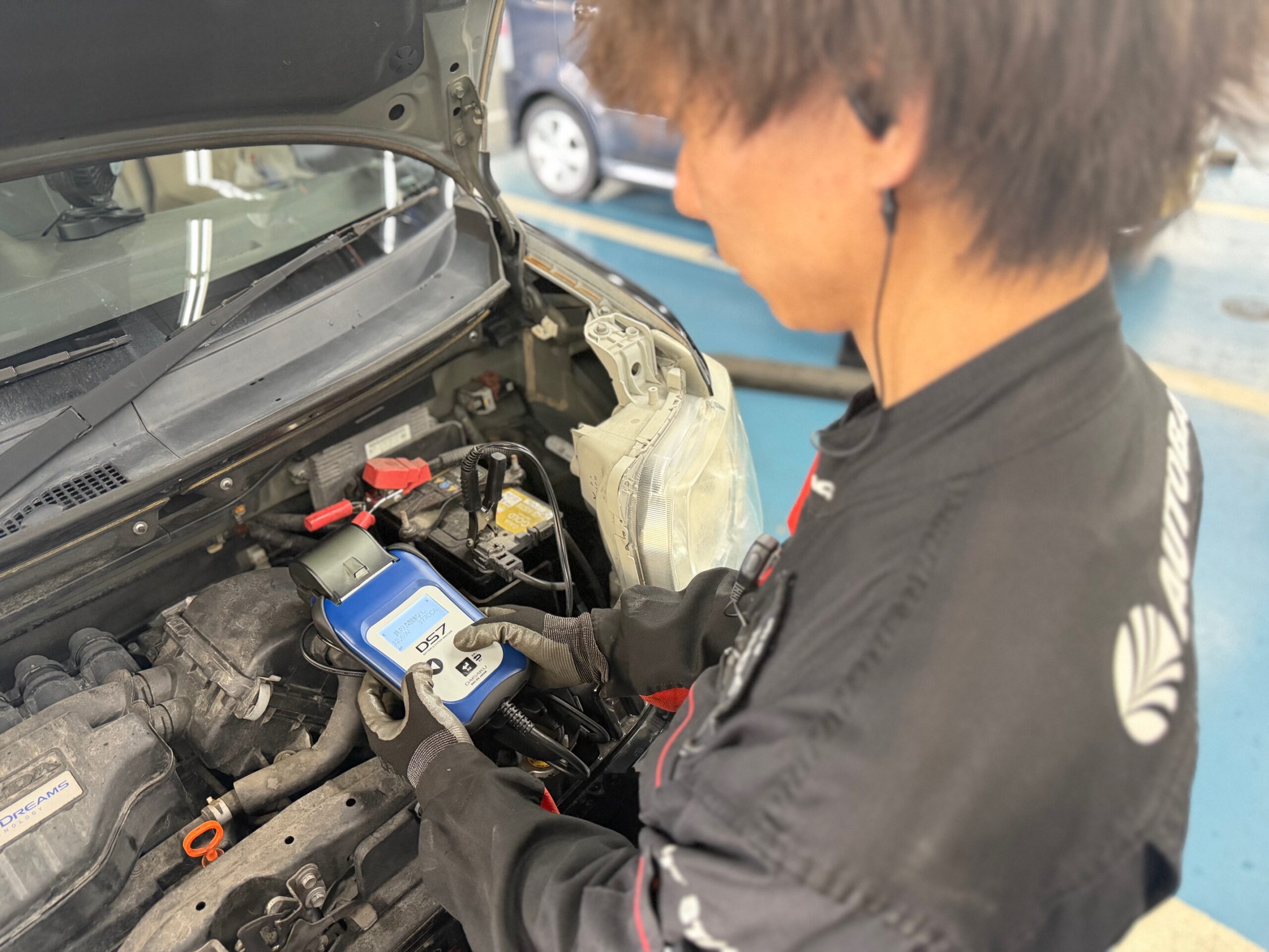 スーパーオートバックスの広いピット内で、ベテラン整備士が懐中電灯を使いながらエンジンルームを細かく、かつ丁寧に見守るように点検している様子