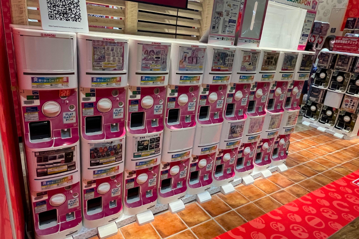 ガシャポンを取り出し、カプセルを開ける瞬間のワクワクした表情の子供のアップ。背景にはカラフルなカプセルの回収ボックスや、たくさんのマシンがぼんやりと映り、ガシャポン体験の楽しさが伝わるイメージ写真