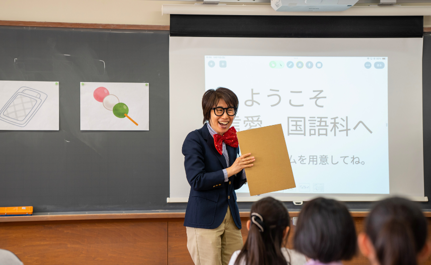 赤い蝶ネクタイをつけた先生が笑顔で国語の授業を進めている様子